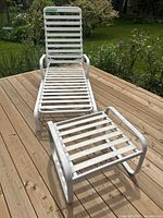 White metal outdoor lounger chair with plastic strap seat and back shown on wooden deck with garden background. Includes matching stool with same design.
