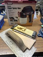 Electric paint spray gun in box with wooden-handled sanding block and yellow hand sander placed in front