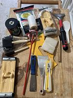 Multiple painting tools arranged on wood surface showing scrapers, brushes, paint mixer, trays, and accessories