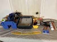 Overview of lot showing blue plastic toolbox organizers with paint brushes, black and white paint trays, yellow drop sheet tool, and other painting tools and rollers arranged on a surface.