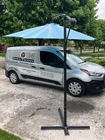 Full view of the large blue offset patio umbrella with black metal frame and cross-shaped base, standing outside on pavement beside a van.