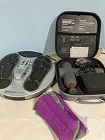 Circulation Booster foot massager, percussion massager in carrying case, and purple ankle weights displayed together on surface