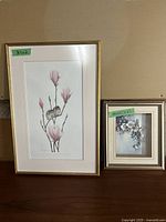Two framed flower pictures side by side against a beige wall and wooden surface, showing size labels.