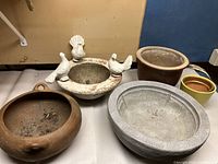 Wide view of resin birdbath top and four various planters on a table, showing condition and size relative to one another.