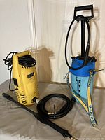 Yellow electric pressure washer and blue Tecnoma sprayer side by side on a gray surface against a light wall.