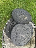 Seven black circular rubber garden stepping tiles stacked on top of one another on a patch of grass and some stone surface, showing the leaf pattern embossing on the top surfaces.