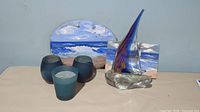 Lot showing 12-inch glass sailboat sculpture with blue sail, oval hand painted beach scene slate wall hanging behind, smaller hand painted sailboat picture, and 3 dark turquoise glass candle holders.