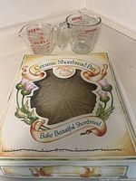 Ceramic Shortbread Pan in its decorative box alongside two Pyrex measuring cups (one smaller, one larger).