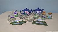 Full lot showing two hand painted ceramic teapots, glass tea cups, three jars, glass pitcher, bowl, covered dish, and two painted stone pieces.