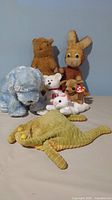 Front group photo of all 7 plush toys arranged facing forward on a beige surface with a blue background wall, showing 4 TY Beanie Babies and 3 other plush toys including a flat frog beanbag.