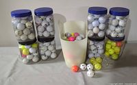 Full view of six clear plastic containers with blue lids filled with used white golf balls, a white plastic basket holding additional golf balls including pink ones, and scattered yellow and white wiffle balls in front.