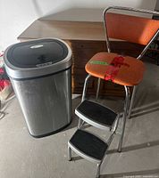 Wide view of orange kitchen stool and stainless steel garbage can side by side on concrete floor with wooden furniture behind.