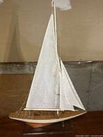 Full view of the vintage model sailboat showing the entire wooden hull, white sails, rigging and wooden stand, in a basement setting.