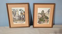 Two vintage Quebec lithographs in matching frames, one missing glass, on display surface.