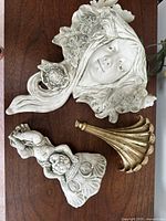Three decorative items: large cement planter wall sconce with female face surrounded by flowers, smaller cherub cement wall planter, and gold colored decorative shelf with ridged pattern.