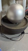Silver plastic ball water fountain sitting inside a black bowl base with power cord on the floor in a basement setting.