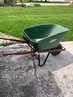 Left side angle of the green Yardworks plastic wheelbarrow showing brown handle frame and black air-filled tire on concrete.