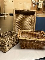 Three items: two large woven wicker baskets and one low rectangular wicker tray placed upright and flat on a surface, against a beige wall.