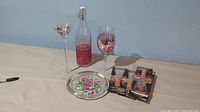 Full view of lot with decorative wine glass, flip-top bottle with pink liquid inside, hand painted plate with floral design, tall hand-painted tea light holder, and four packages of LED bottle lights.