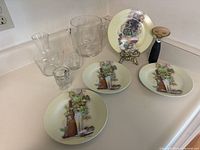 Photo showing four small wine-themed ceramic plates arranged with various glassware including a whisky decanter, a glass ice bucket, a shot glass, a corkscrew, and a brass plate holder stand.
