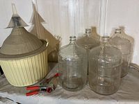 Overview of four large clear glass wine jugs, a large plastic wine barrel, and a double lever hand corker with red handles placed on a white surface against a wall.