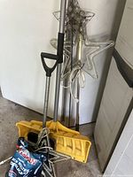 Two yellow snow shovels with metal handles, two star-shaped lawn ornaments with cords for lighting, leaning against a wall indoors.