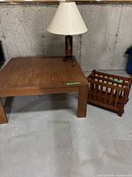 Wooden coffee table, wooden lamp with beige shade, and wooden magazine holder placed on concrete floor against concrete wall