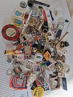 Overview of assorted collectible items including keychains, pins, badges, pipe, pocket watch, spoons, and branded collectibles spread on white fabric.