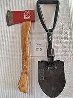 Photo of a used axe with a wooden handle and red painted head, next to a black folding shovel with a triangular handle and serrated blade, items placed on a white textured surface.
