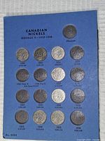 Blue cardstock holder page labeled 'Canadian Nickels George V 1910-1936' containing seven silver-colored Canadian 5-cent coins from 1922 to 1935 inserted in cutouts.