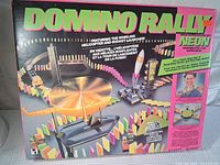 Full front of the Domino Rally game box showing branding, game setup with dominoes, helicopter, and rocket launcher on front cover.