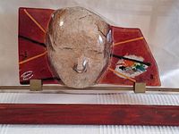 Front view of the handmade glass artwork showing the abstract mask face and red textured background mounted on a wood frame.