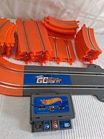 Photo showing various orange straight and curved track pieces alongside the grey base track piece with Go For It! branding, and the electronic control unit placed on a white cloth.