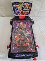 Full view of Power Rangers pinball toy on a white surface, showing colorful playfield with Red, Black and Yellow Rangers and mechanical metal bumpers, flippers, and clear plastic top cover.