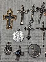 Close-up photo of assorted religious pendants featuring various crucifixes and round/oval medallions laid on fabric background.