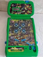 Closed and open views of a tabletop pinball game with Teenage Mutant Ninja Turtles artwork on the playfield and the scoreboard cover.
