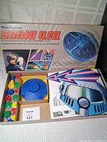 Photo showing the open game box containing the blue electronic UFO game unit, colorful game pieces, and the box lid with the game title and art.