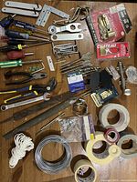 Photo showing assorted hand tools including pliers, hand saw, various screws, tape rolls, metal wires, and packaged hardware items.