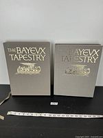 Closed view of the Bayeux Tapestry coffee table book and matching slipcase, both gray with gold lettering and illustration.