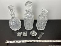 Three clear crystal bottles with cut and etched designs, accompanied by several small crystal dishes and lids. Tape measure shows scale.