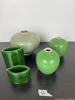 Front view of five green glazed ceramic pieces: one flattened ovoid vase, two round bud vases, two small leaf-shaped vases.