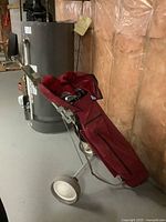 Folding two-wheel metal golf cart with attached red nylon golf bag leaning against basement wall near water heater.