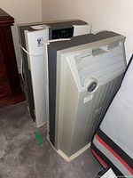 Two standing white and gray Nikken air purifiers positioned side by side on carpeted floor next to a bed and wooden furniture.