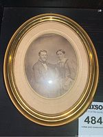 Oval antique portrait of a couple seated, black and white photo inside a gold round frame with glass and mat, viewed from front.