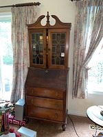 Full view of the antique secretary desk standing between two windows with drawn floral curtains showing the entire piece including the glass door cabinet on top and three drawers below the slant-top desk.
