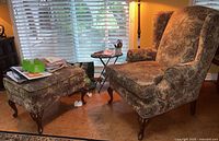 Wingback armchair with ottoman placed near window, showing both items and some clutter on ottoman