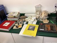 Photo showing books, paper pads, white decorative moulding pieces, bags of shells, stamps and colored pencils on a green surface.
