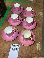 Six pink Aynsley demitasse cups on matching saucers arranged on a wooden surface, some with visible gold trim