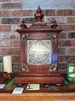 Front view of wooden Westminster chime bracket clock on shelf with three winding keys in front, showing decorative brass corner accents and finial top.