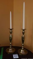 Two solid brass candlesticks with white taper candles shown side by side on a table against a wall